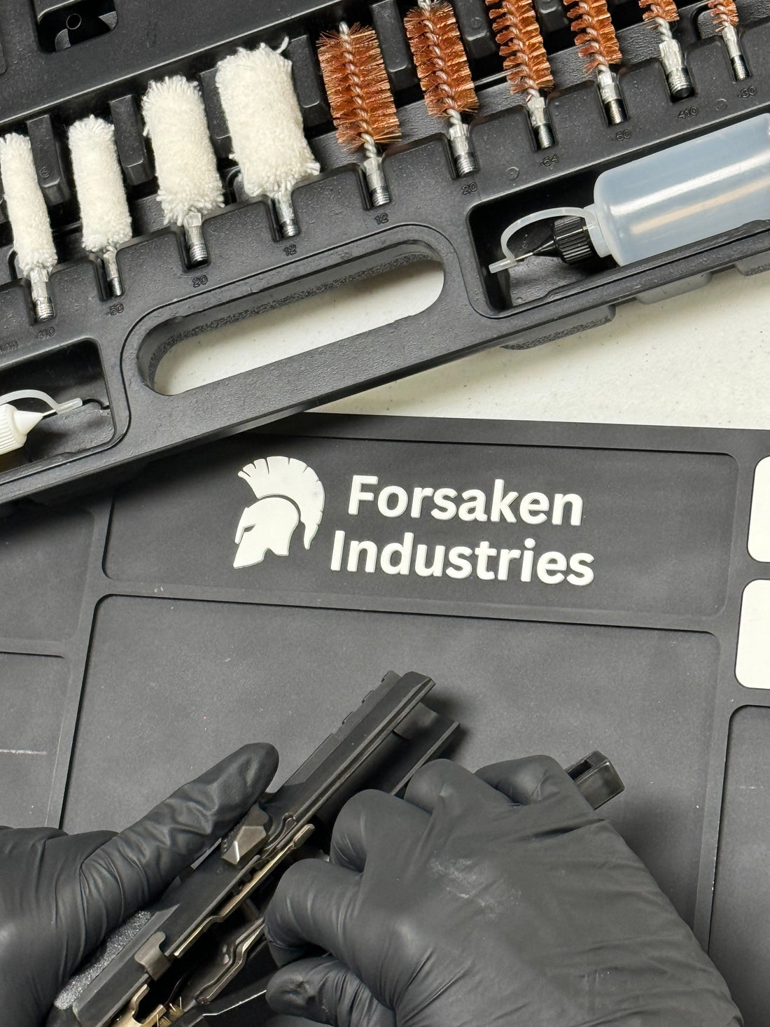 Person cleaning a disassembled pistol on a magnetic gun cleaning mat with CLP and carbon cleaner bottles on the table