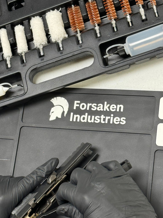 Person cleaning a disassembled pistol on a magnetic gun cleaning mat with CLP and carbon cleaner bottles on the table