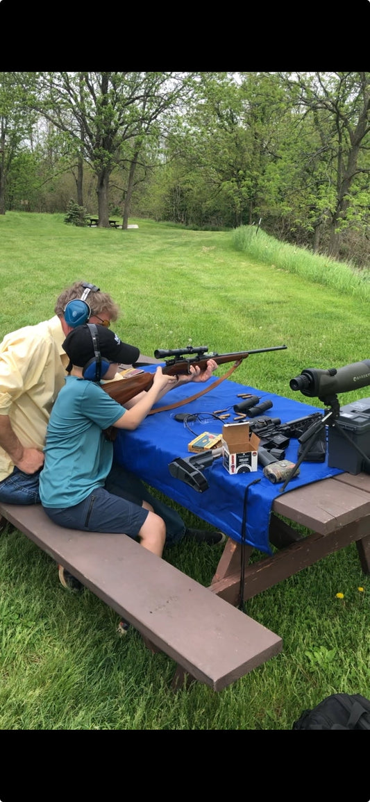 Father teaching son how to shoot at an outdoor range, both wearing eye and ear protection, focused on firearm safety and technique